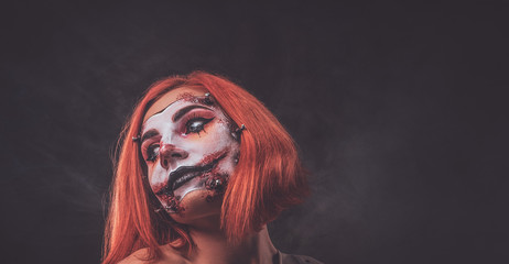 Pretty young girl with scary dead doll makeup is posing for photographer at dark photo studio.