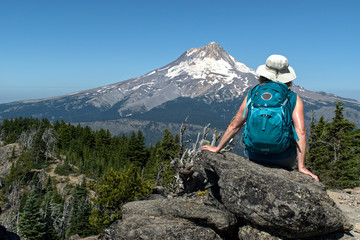 A mature woman hiker sites on a rock with her back to us and enjoys the view of Mt. Hood from Lookout Mt. There is a clear blue sky and bright summer sunlight.