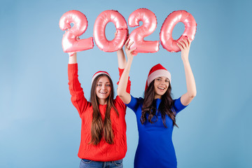 Wall Mural - two smiling women wearing santa hats holding 2020 balloons for new year holiday isolated over blue