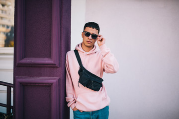 Wall Mural - City portrait of handsome hipster guy wearing pink blank hoodie with black waist bag with space for your logo or design. Mockup for print