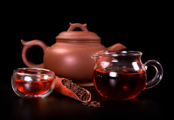 Wall Mural - Ceramic tea pot, glass jug with cup of tea and dried black tea in a scoop on a black background.