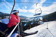 Happy Female Tourist Is Sitting On Chairlift In Mountain Resorts. Girl In A Ski Suit At An Altitude And Shows With Hand On Snow-capped Mountains