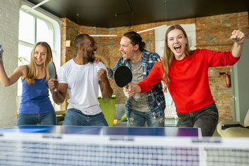 Wall Mural - Young people playing table tennis in workplace, having fun. Friends in casual clothes play ping pong together at sunny day. Concept of leisure activity, sport, friendship, teambuilding, teamwork.