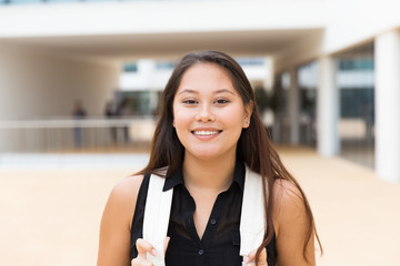 Wall Mural - Happy excited female tourist posing outside. Young mix raced woman in casual standing near city building, holding backpack straps, looking at camera, smiling. Young woman portrait concept