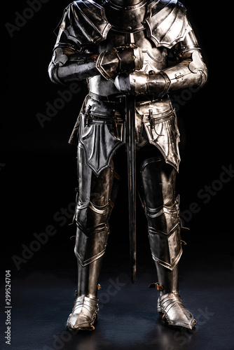 Fotomural cropped view of knight in armor holding sword on black background
