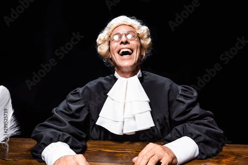 Fotomural smiling judge in judicial robe and wig sitting at table isolated on black