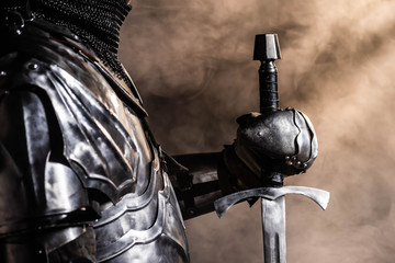 cropped view of knight in armor holding sword on black background