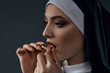 Horizontal half-turn close-up portrait of a nun, posing on a black background. She wearing dark nun's clothing. The nun brings a cigarette to her mouth and licking it.  