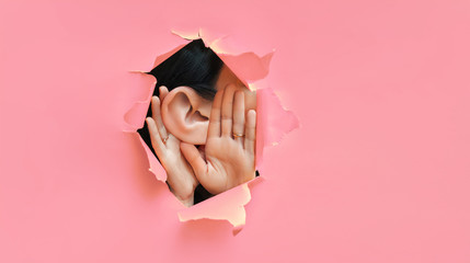 Wall Mural - Female ear and hands close-up. Copy space. Torn paper, pink background. The concept of eavesdropping, espionage, gossip and the yellow press. Caricature with an enlarged ear.