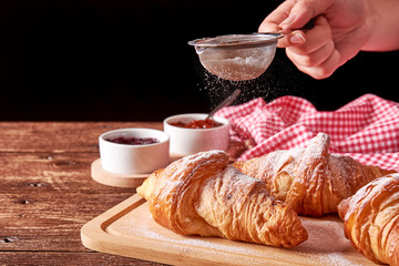 Still life on old wooden mat with three sugared croissants and fruit jam bowls. Hand holding a strainer of sugar