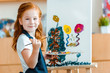 © LIGHTFIELD STUDIOS - smiling redhead child standing near painting on canvas in art school