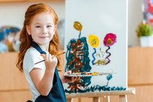 Smiling Redhead Child Standing Near Painting On Canvas In Art School