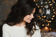 Close up portrait of brunette model girl posing in an interior with festive Christmas lights on the background. Young beautiful smiling woman with a trendy makeup and smooth skin looking aside