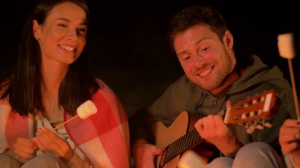 Poster - leisure and people concept - group of smiling friends sitting at camp fire, roasting marshmallow and playing guitar at night