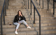 © Anastasia Rudenko - Portrait of european young beautiful smiling woman with dark straight hair sitting on stairs in black leather jacket and blue jeans with drink in hand