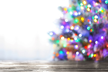 Blurred view of beautiful Christmas tree with colorful lights near window indoors, focus on wooden table. Space for text