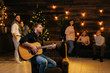 © dikushin - Handsome cheerful man plays guitar and sings in front of friends at a Christmas party in a cozy house. Guys and girls singing to their friend. Christmas tree with garland in background.