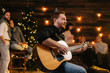 © dikushin - Portrait of young happy man in the festive hat singing with guitar. against the background of talking friends. Christmas tree with garland and wall with festive illumination in background.
