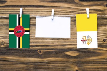 Hanging flags of Dominica and Vatican City attached to rope with clothes pins with copy space on white note paper on wooden background.Diplomatic relations between countries.