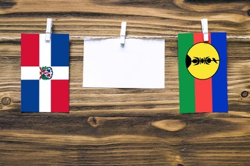 Hanging flags of Dominican Republic and New Caledonia attached to rope with clothes pins with copy space on white note paper on wooden background.Diplomatic relations between countries.