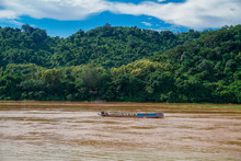 A Traditional Laos Slow Boat Cruising A Muddy Brown Lower Basin Mekong River After High Rainfall. Tropical Jungle Covers The Mountainous Background Of The UNESCO World Heritage Site Of Luang Prabang