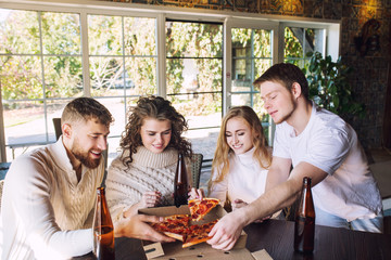 Friends four two men and two women young adults beautiful and happy together at the table eating pizza and drinking drinks from bottles