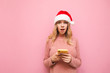 Shocked girl in christmas hat holds smartphone and looks surprised at camera on pink background. Surprised beautiful girl in pink sweater, uses smartphone, isolated. Xmas concept