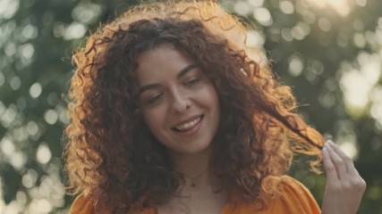 Sticker - Beautiful young redhead woman in orange dress smiling and playing with her curly hair while resting in the park