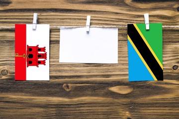 Hanging flags of Gibraltar and Tanzania attached to rope with clothes pins with copy space on white note paper on wooden background.Diplomatic relations between countries.