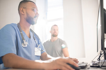 Serious doctor filling in electronic document with data of patient by computer