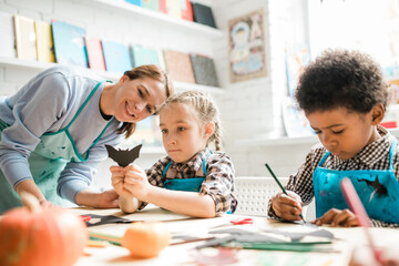 Poster - One of multicultural schoolkids showing handmade black paper bat to teacher
