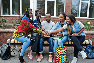 Group of five african college students spending time together on campus at university yard. Black afro friends studying at bench with school items, laptops notebooks.