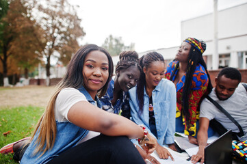 Group of five african college students spending time together on campus at university yard. Black afro friends sitting on grass and studying with laptops.