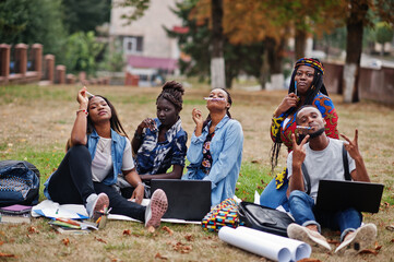 Wall Mural - Group of five african college students spending time together on campus at university yard. Black afro friends sitting on grass and studying with laptops.