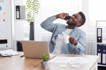 Poster - Joyful african businessman talking on phone and drinking coffee at workplace