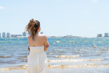 Wall Mural - Young woman back cold shivering standing in white dress on beach bay shore in Florida with cityscape skyline during day of Pensacola