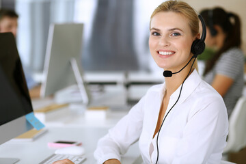 Blonde business woman using headset for communication and consulting people at customer service office. Call center. Group of operators at work at the background