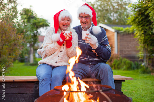 Cuadros en Lienzo Senior couple with Christmas santa's hats and hot drinks in front of a fireplace