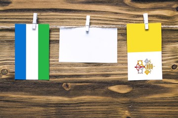 Hanging flags of Sierra Leone and Vatican City attached to rope with clothes pins with copy space on white note paper on wooden background.Diplomatic relations between countries.