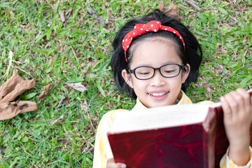 Wall Mural - Cute little girl with glasses lying on grass reading book