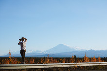 Young Man Standing On Barrier Next To The Highway And Photographing Alaskan Mountains