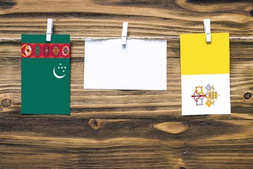Hanging flags of Turkmenistan and Vatican City attached to rope with clothes pins with copy space on white note paper on wooden background.Diplomatic relations between countries.