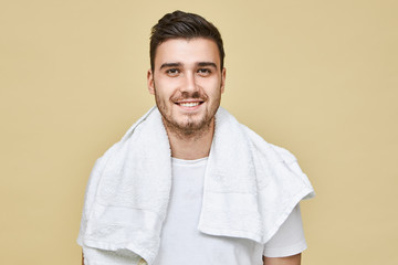 Wall Mural - Studio image of attractive cheerful young European male with stubble and white towel around his neck smiling broadly at camera, going to shave his face in bathroom in the morning before work