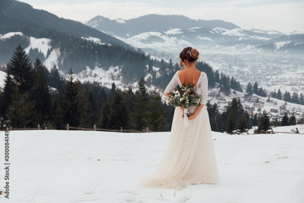 Bride in white wedding dress holding colorful flowers bouquet in hands and posing outdoors. Winter wedding and season floral concept. Mountains on background. - obrazy, fototapety, plakaty 