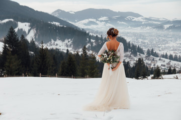 Bride in white wedding dress holding colorful flowers bouquet in hands and posing outdoors. Winter wedding and season floral concept. Mountains on background.