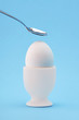 One white egg in an egg cup with a teaspoon hitting the egg shell on blue background