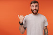 Happy pretty bearded tattooed man with trendy haircut looking at camera with wide smile and rounding his blue eyes, pointing aside with raised thumb while posing over orange background