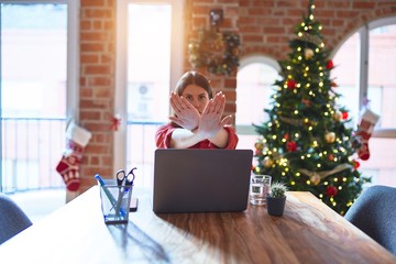 Poster - Beautiful woman sitting at the table working with laptop at home around christmas tree Rejection expression crossing arms and palms doing negative sign, angry face