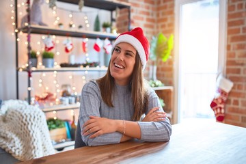 Sticker - Young beautiful woman wearing christmas hat sitting at the table at home looking away to side with smile on face, natural expression. Laughing confident.