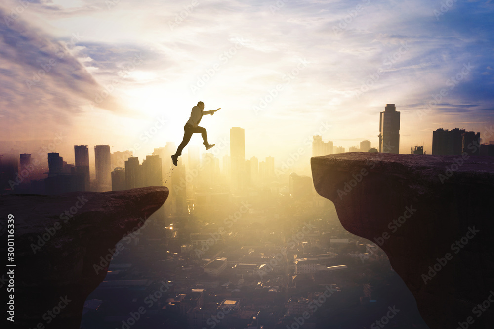 Businessman jumping through gap cliff at sunrise - obrazy, fototapety, plakaty 